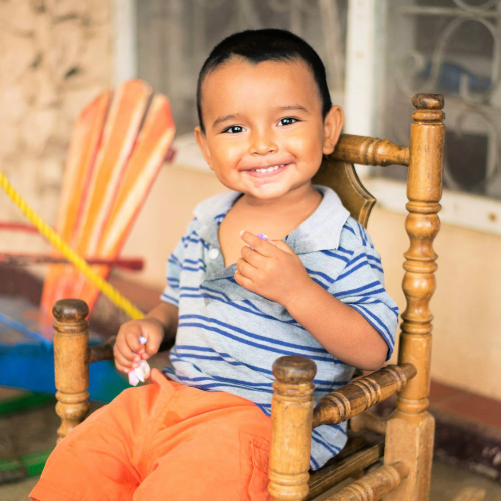 A young boy with dark hair a gray and blue striped shirt sits on a wooden chair with a huge smile on his face.