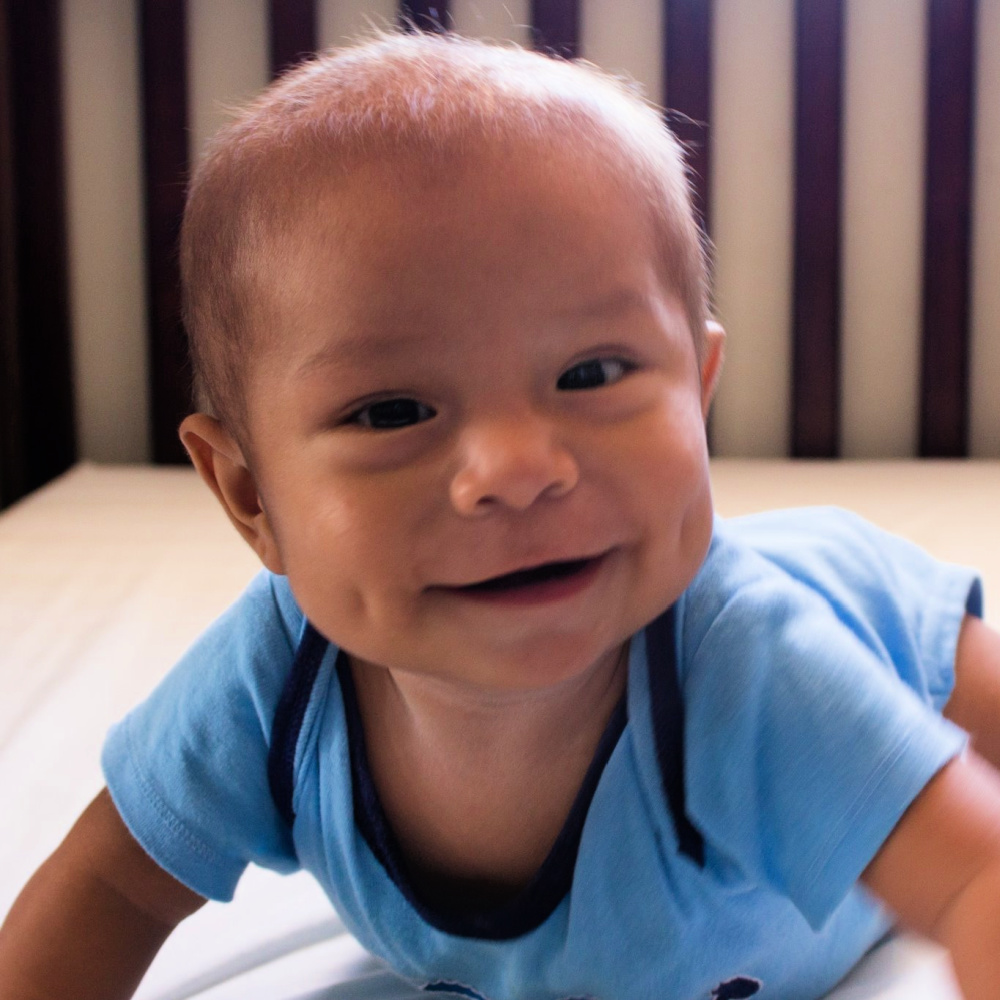 A baby with dimples, light brown hair, and a blue onesie smiles at the camera.