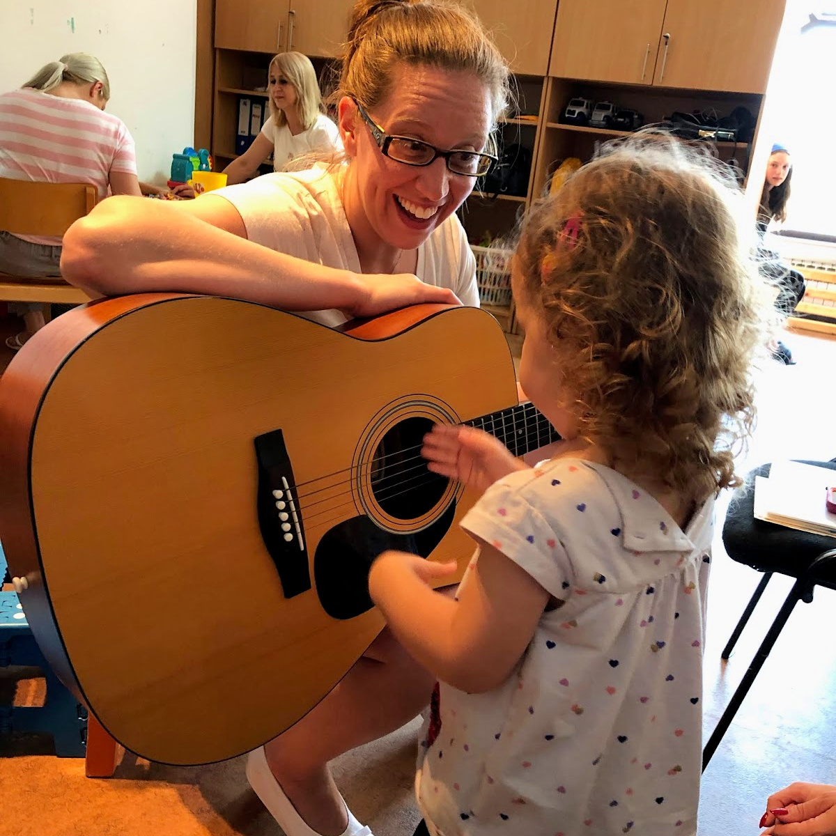 A young woman with a guitar smiles and invites a young girl to play and strum the strings.