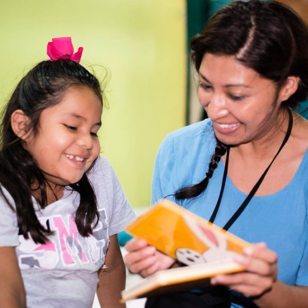 A young girl and a caregiver sitting close together with smiles on their faces while reading a book.