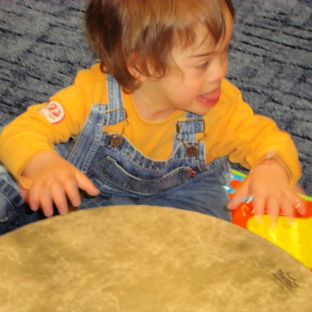A young boy with Down syndrome, sitting on the floor, playing a gathering drum.