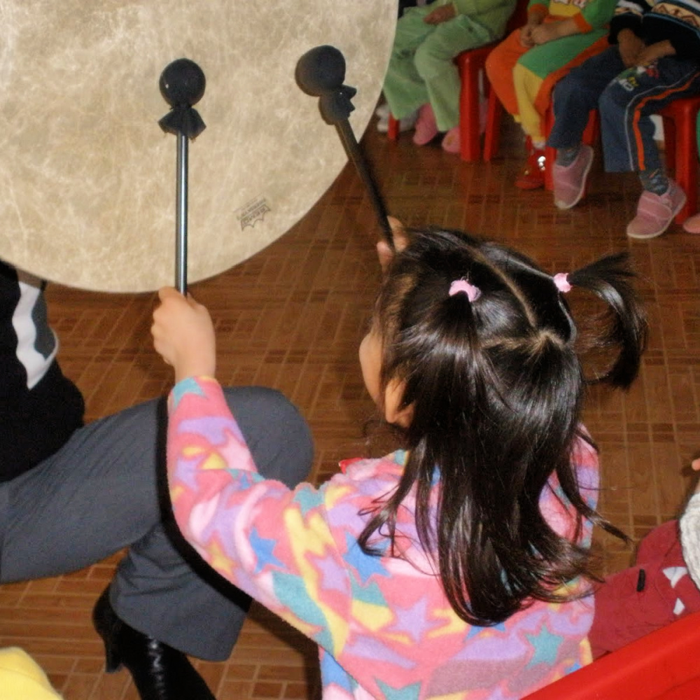 A child with dark hair holding two drum mallets, playing happily on a gathering drum.