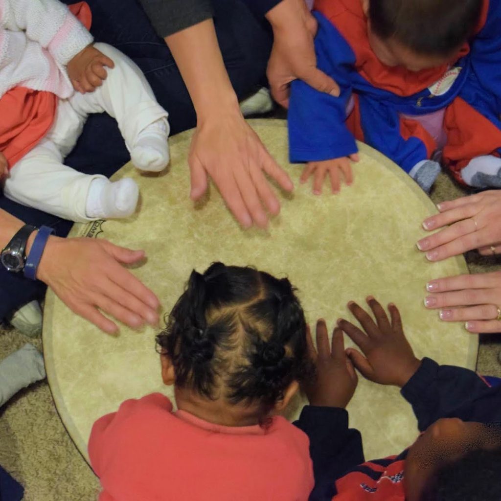 An overhead view of several babies and toddlers drumming together with their hands on a gathering drum.