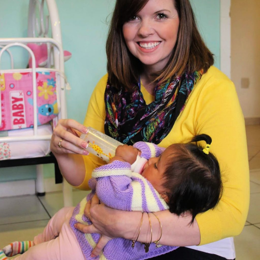 A woman in a bright yellow sweater and colorful scarf feeding a bottle to a very young girl.