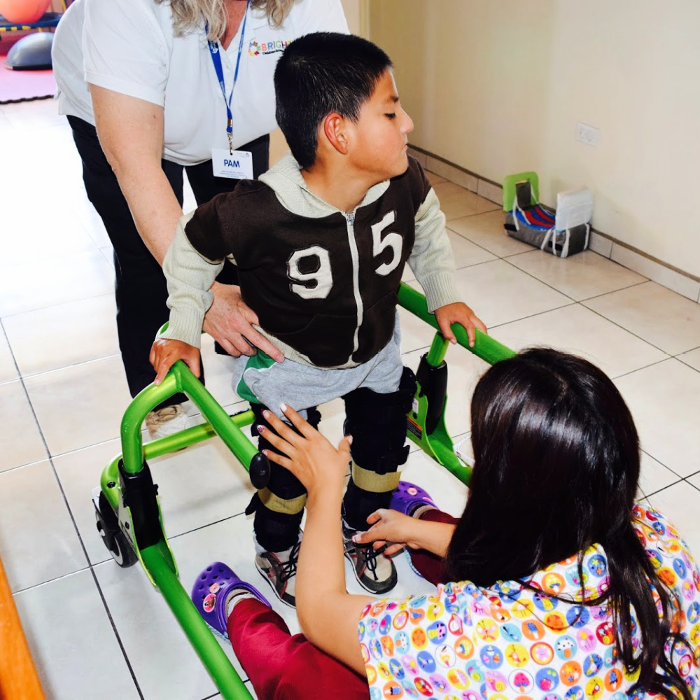 A young boy standing with the assistance of 2 therapists and a bright green walking assistive device.