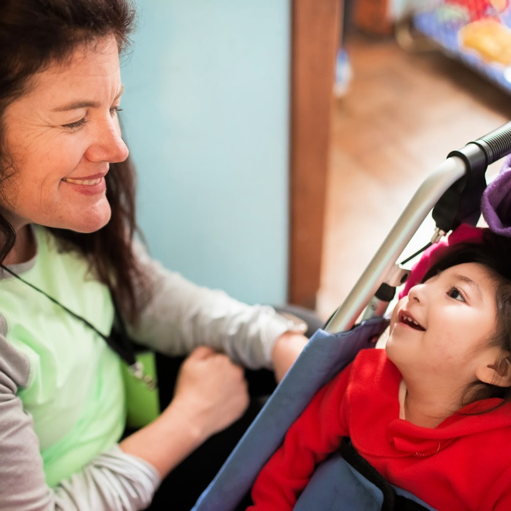 A young child in a wheelchair looking up at an adult caregiver with a smile on her face.