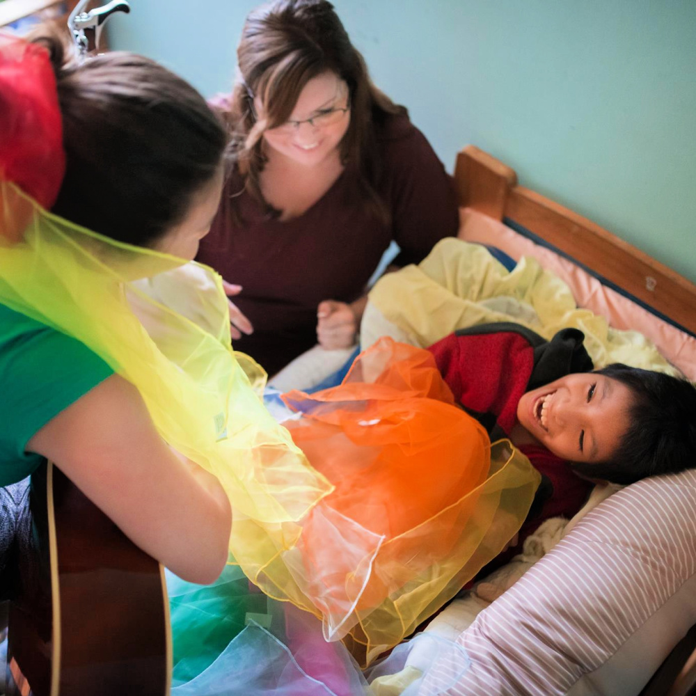 A young person laying in bed with a smile on their face. Caregivers and a music therapist surround the bed, which is covered in colorful movement scarves.